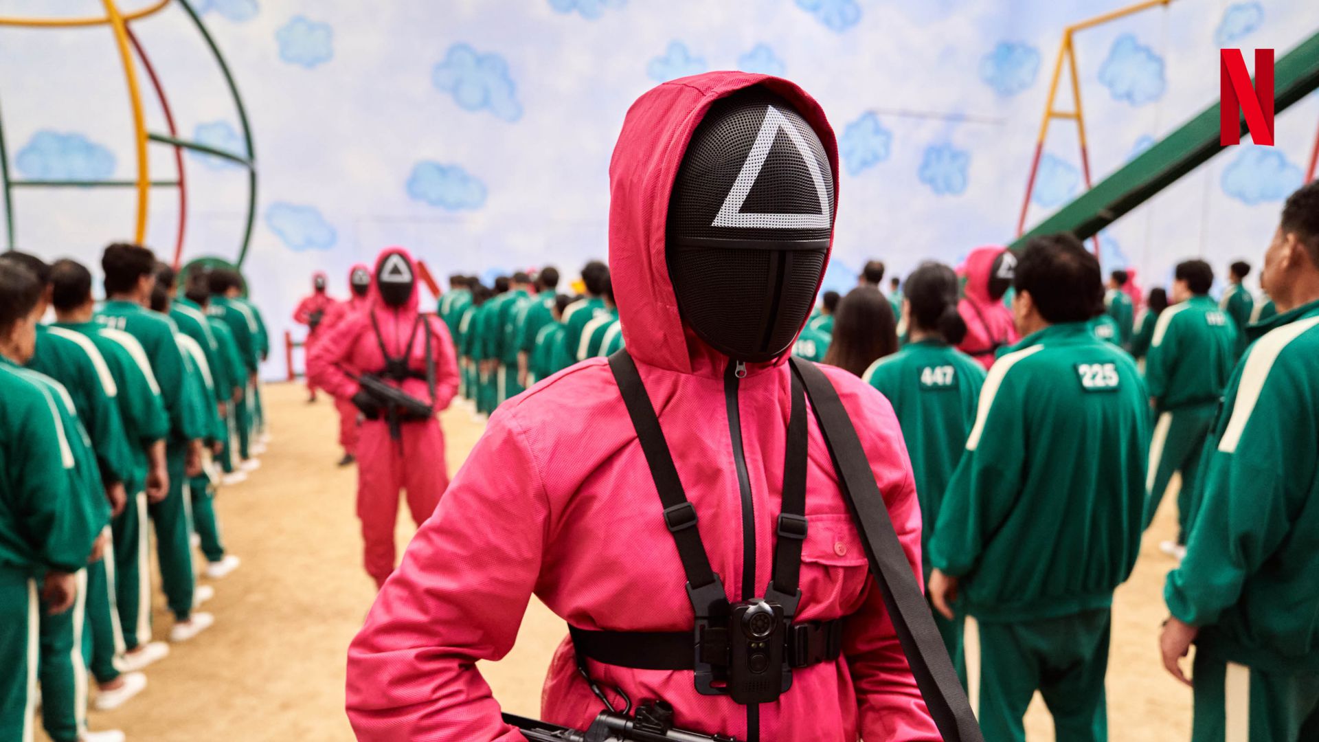 Una guardia con una tuta rossa con cappuccio e una maschera nera con un triangolo si trova di fronte a concorrenti che indossano tute da ginnastica verdi, in un'ampia area interna con attrezzature da gioco. Il logo di Netflix appare nell'angolo in alto a destra.