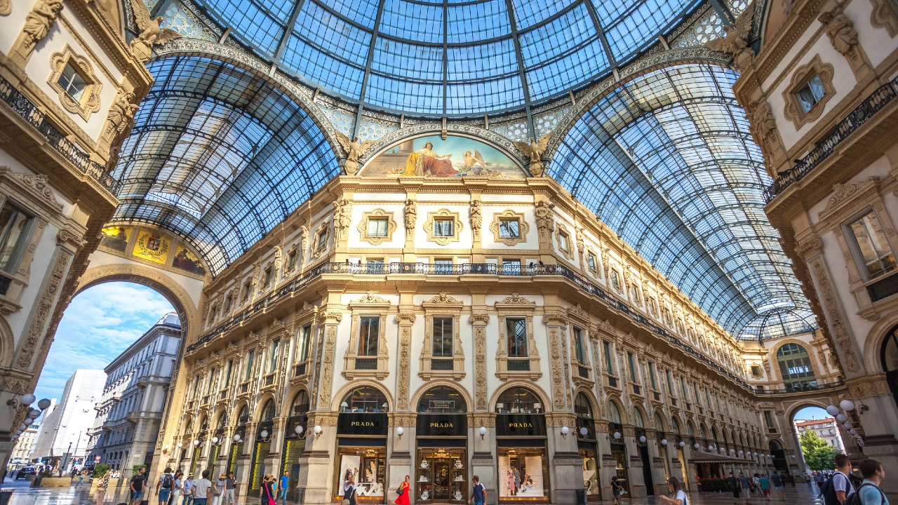 L'elegante interno della Galleria Vittorio Emanuele II di Milano, un tempo nota come Mediolanum, è caratterizzato da un'architettura ornata, soffitti ad arco in vetro, affreschi decorativi e vetrine di lusso con persone che camminano sotto la luce del sole.