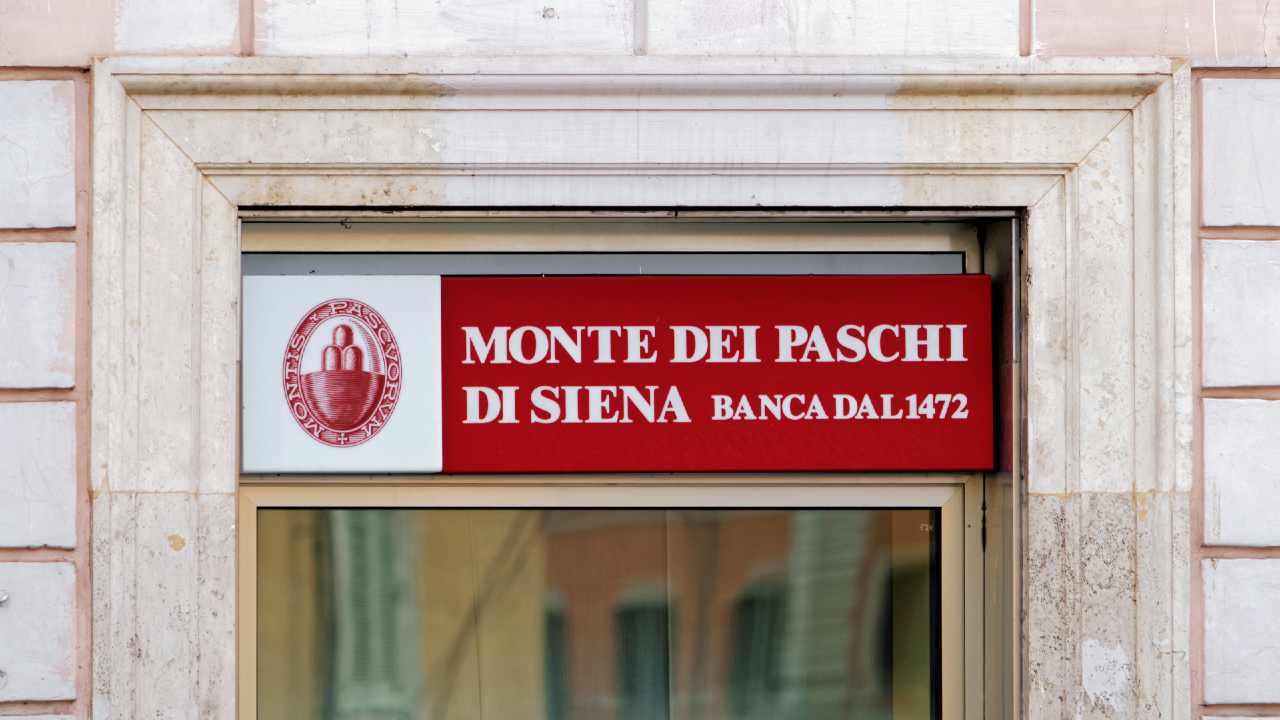 Un'insegna sopra un portone recita "Monte dei Paschi di Siena Banca dal 1472" con un logo sulla sinistra, incastonato nella facciata di un edificio in pietra.
