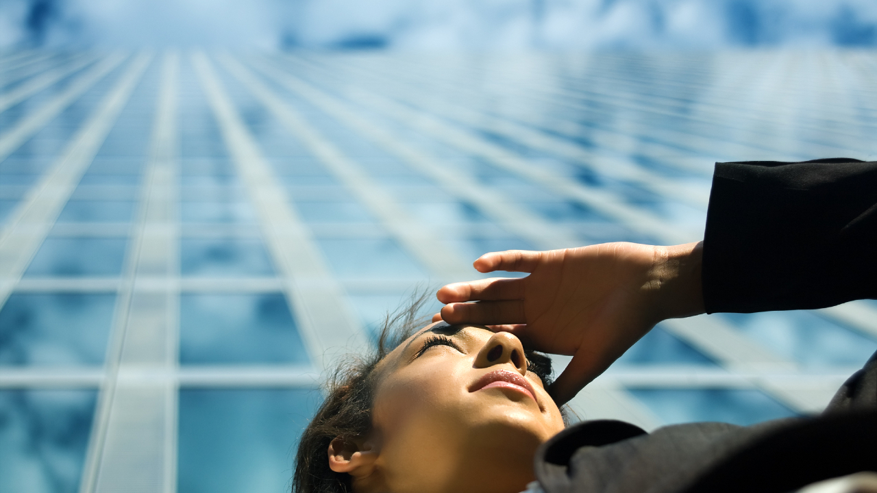 A person in business attire looks up, shading their eyes with one hand, against the backdrop of a tall glass building reaching into a cloudy sky. The perspective is from below, emphasizing the building’s height.