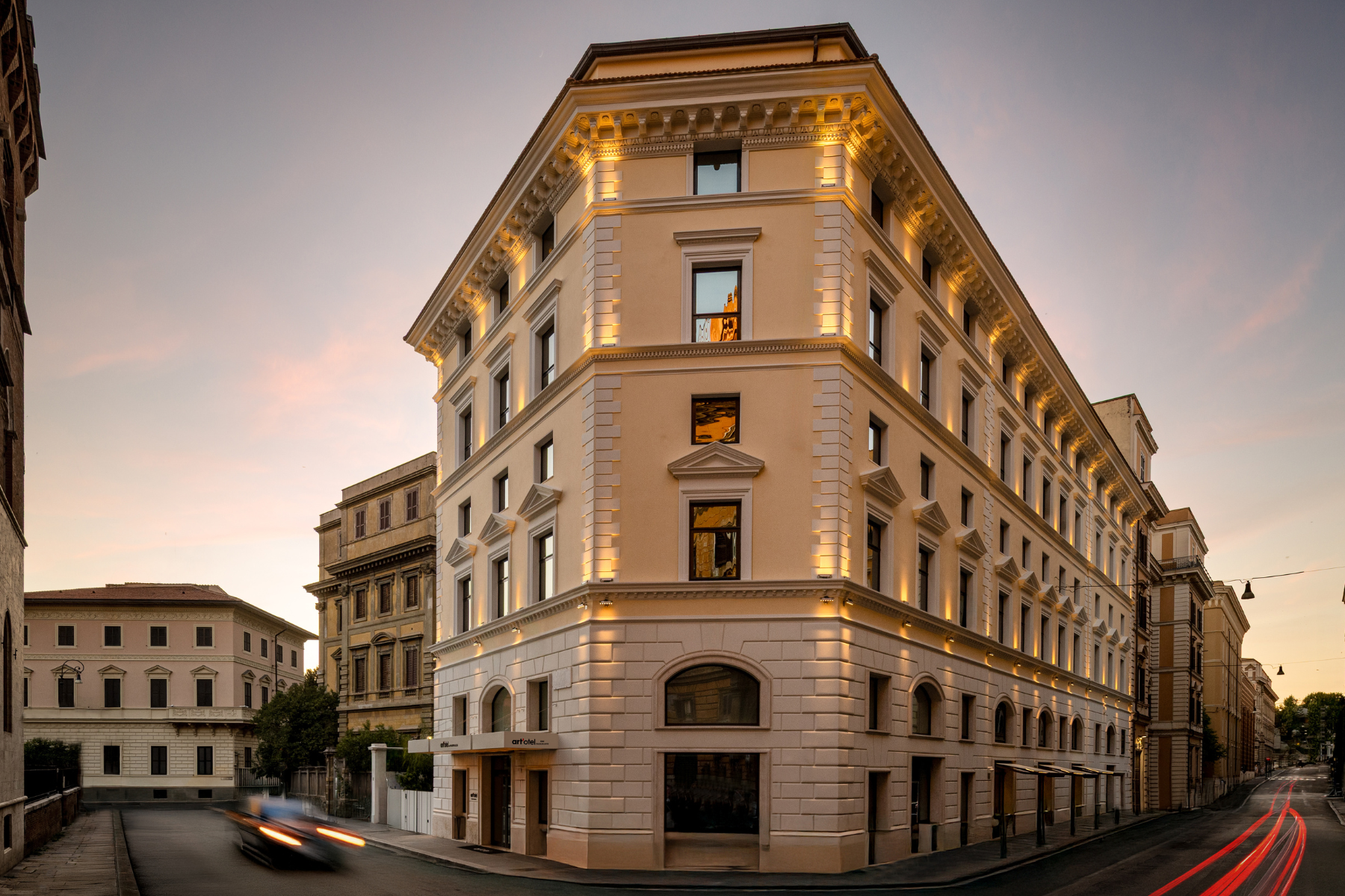 A grand, beige stone building with illuminated windows and ornate architectural details stands on a street corner at dusk, with light trails from passing cars visible in the foreground.