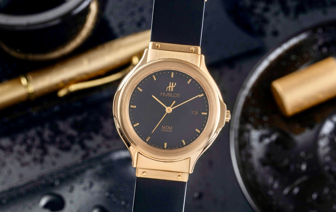 Un orologio da polso Hublot in oro con quadrante e cinturino neri è esposto in bella mostra con gocce d'acqua intorno, una penna d'oro e un piatto sfocato sullo sfondo: una scelta elegante per gli appassionati di orologi. L'orologio indica le 10:08 e la data il 13.