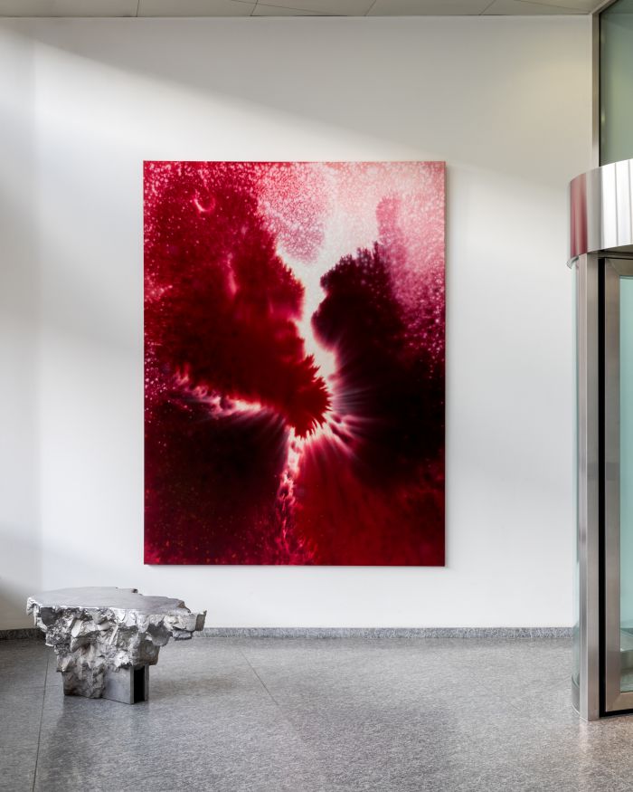Una grande opera d'arte astratta della collezione Julius Baer arte, con vivaci sfumature rosse e rosa, è appesa a una parete bianca in uno spazio moderno. Sotto di essa si trova un tavolo scultoreo in pietra su un pavimento grigio maculato, vicino a una porta in vetro e metallo.