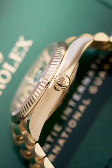 Vista laterale ravvicinata di un orologio Rolex d'oro con lunetta scanalata e corona, appoggiato su una scheda di garanzia Rolex verde. La messa a fuoco evidenzia i dettagli dell'orologio, evocando il fascino e l'intricata maestria della collezione Rolex Puzzle.