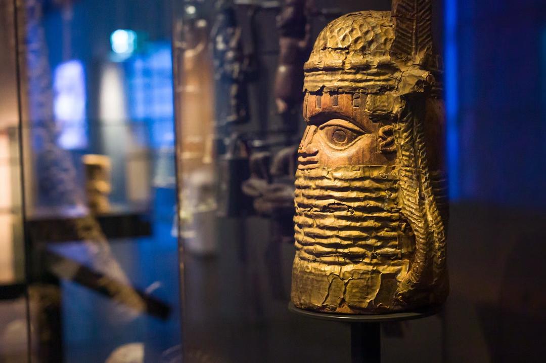 Primo piano di un'antica maschera di legno intagliata con motivi intricati e una barba dettagliata, esposta in una mostra museale poco illuminata. Lo sfondo blu, morbidamente sfocato, evoca l'atmosfera coinvolgente di un videogioco liberare arte.