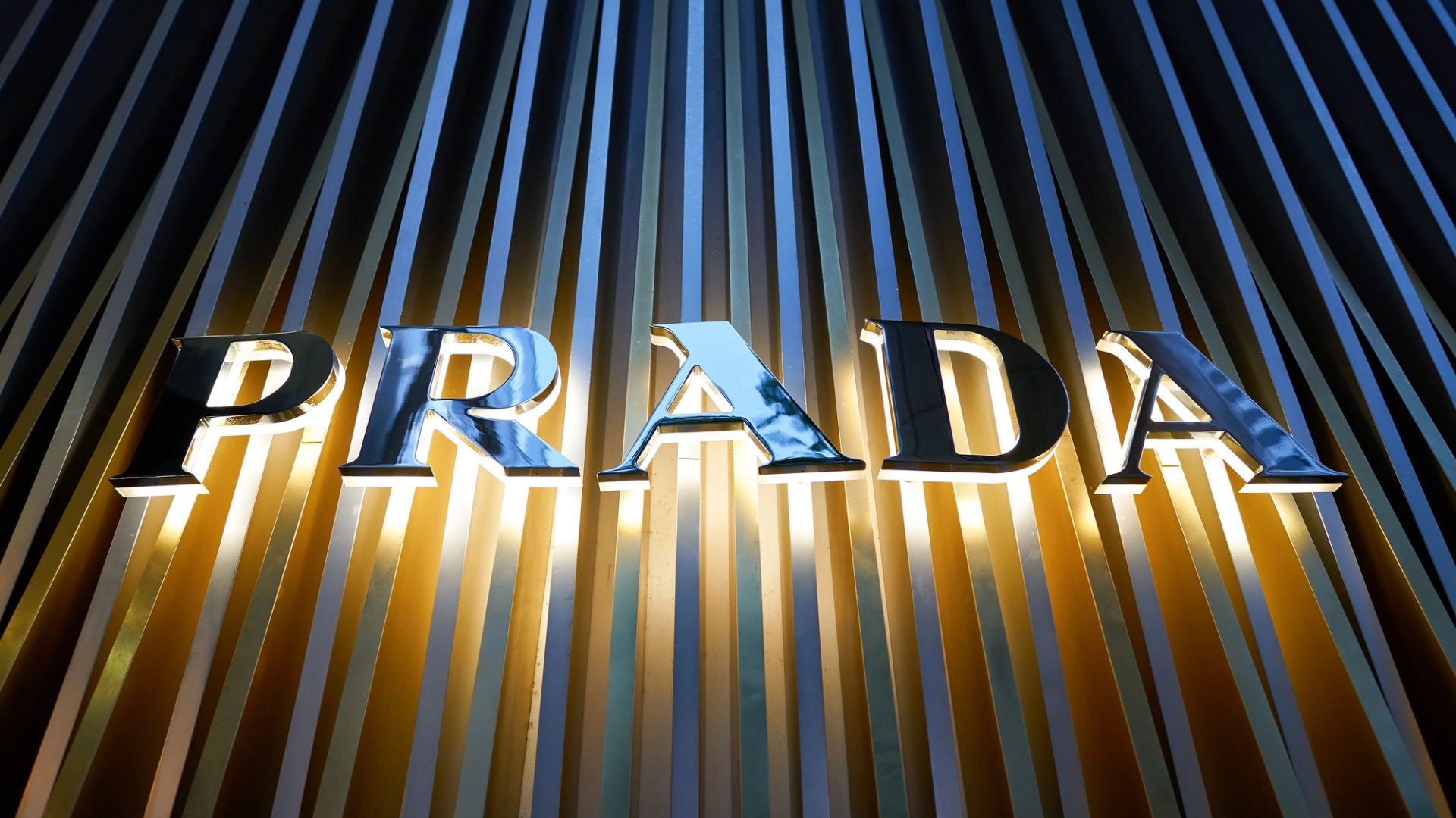 Il logo Prada a lettere metalliche è illuminato su uno sfondo a strisce verticali oro e argento.