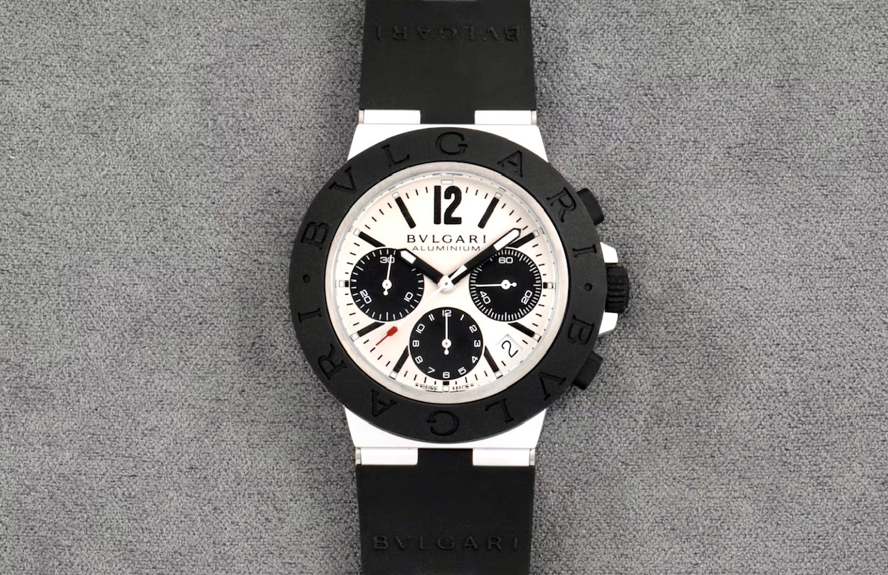 Un orologio da polso BVLGARI in alluminio con cinturino in caucciù nero, quadrante bianco, contatori neri e lunetta nera con inciso "BVLGARI" su sfondo grigio: una scelta ideale per l'estate.