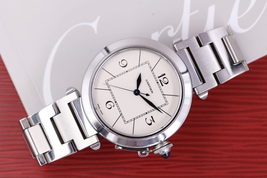 Un orologio da polso Cartier d'argento con quadrante rotondo bianco, numeri neri e cinturino in acciaio inossidabile - un ideale orologio d'epoca - poggia su una superficie rossa strutturata vicino a una scatola bianca con la scritta "Cartier" parzialmente visibile.