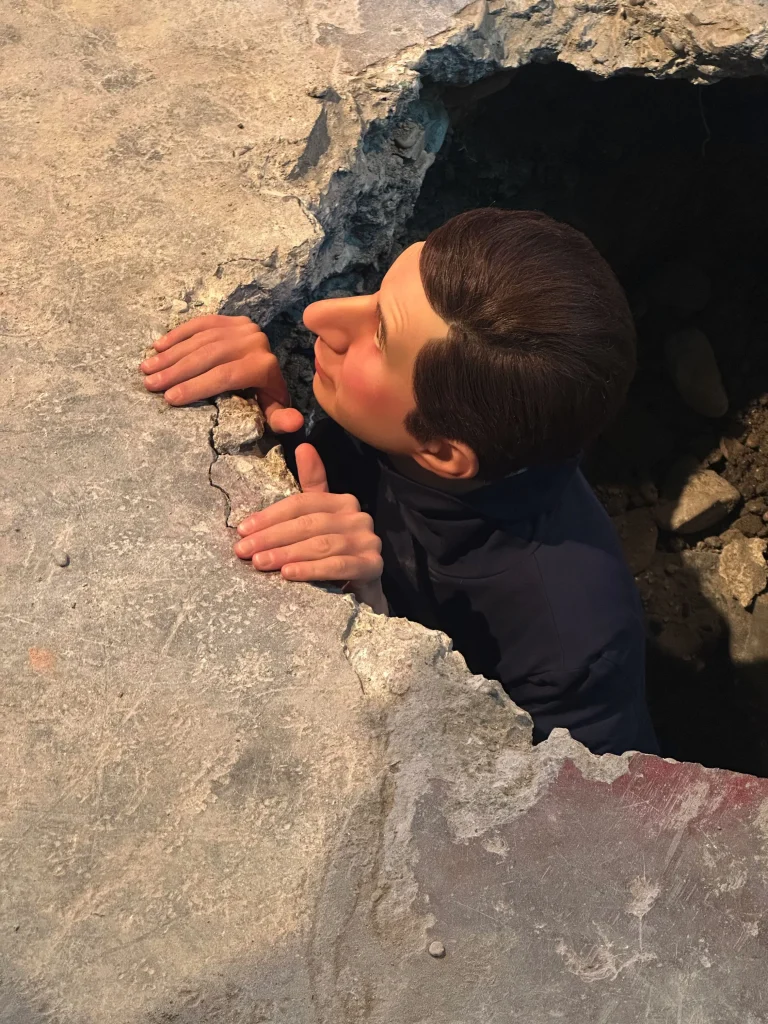 Una persona con i capelli corti e scuri sbircia attraverso un buco in una superficie di cemento crepata, afferrando il bordo con entrambe le mani, ricordando Santa di Maurizio Cattelan e il suo aspetto inaspettato e surreale.