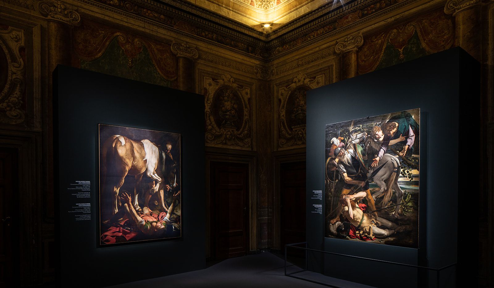 Due grandi dipinti in cornici ornate sono esposti su pareti scure in una sala riccamente decorata con soffitti affrescati e ritratti classici, illuminati da una luce mirata.