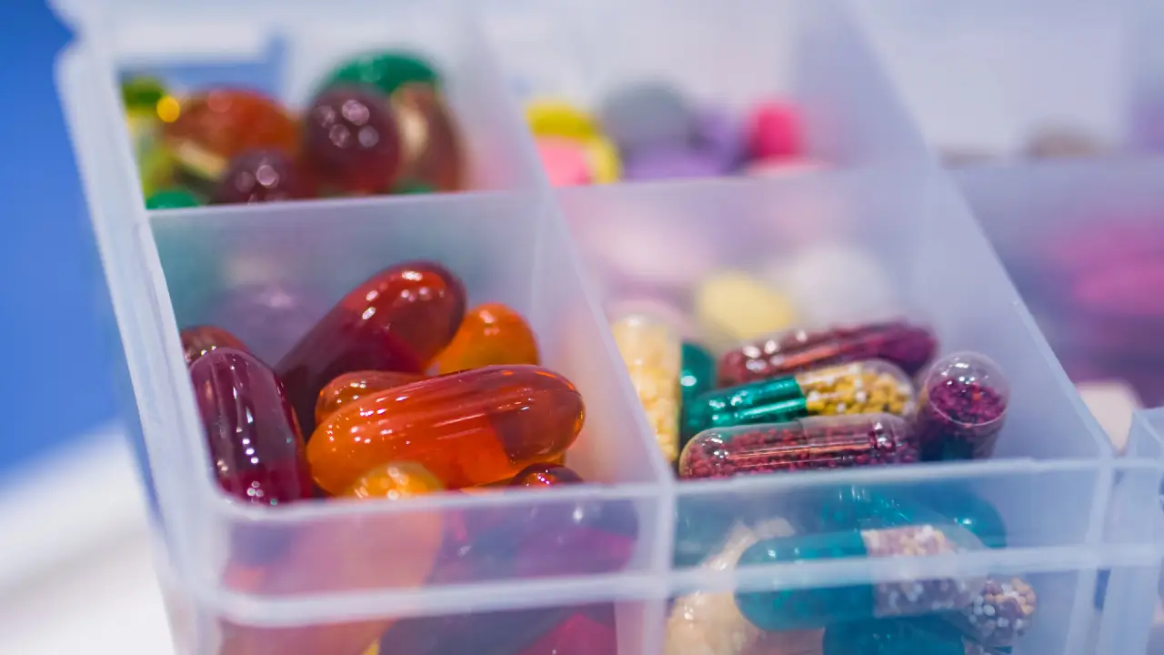 Primo piano di un organizer per pillole in plastica contenente varie capsule e pillole colorate, disposte in scomparti separati. Le pillole sono di forme, dimensioni e colori diversi, tra cui rosso, arancione, giallo, verde e trasparente.