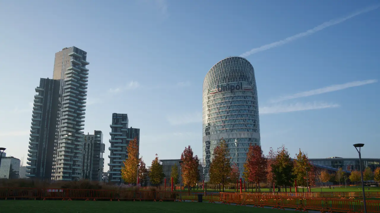 Un paesaggio urbano caratterizzato da moderni grattacieli, tra cui una torre cilindrica denominata "Unipol", con alberi autunnali e un parco erboso in primo piano sotto un cielo azzurro e limpido.