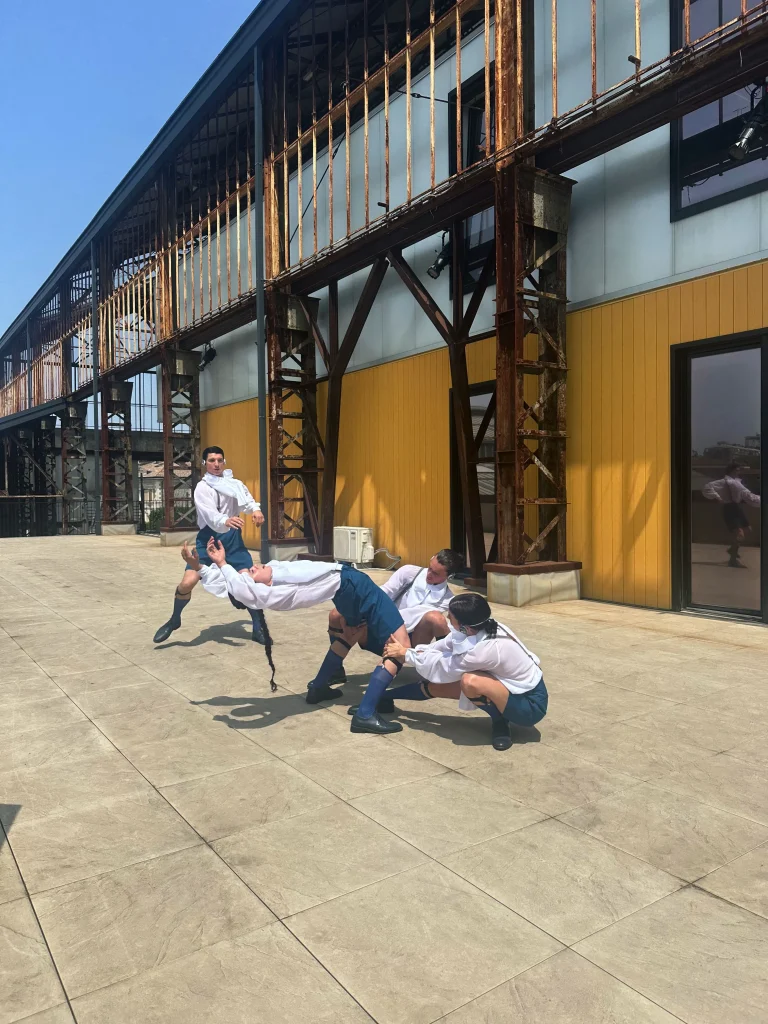 Quattro persone in camicia bianca, pantaloncini marini e calzini blu si esibiscono in una posa dinamica e acrobatica su un'area di cemento soleggiata accanto a un edificio giallo e bianco in stile industriale, evocando lo spirito giocoso di Maurizio Cattelan Santa.