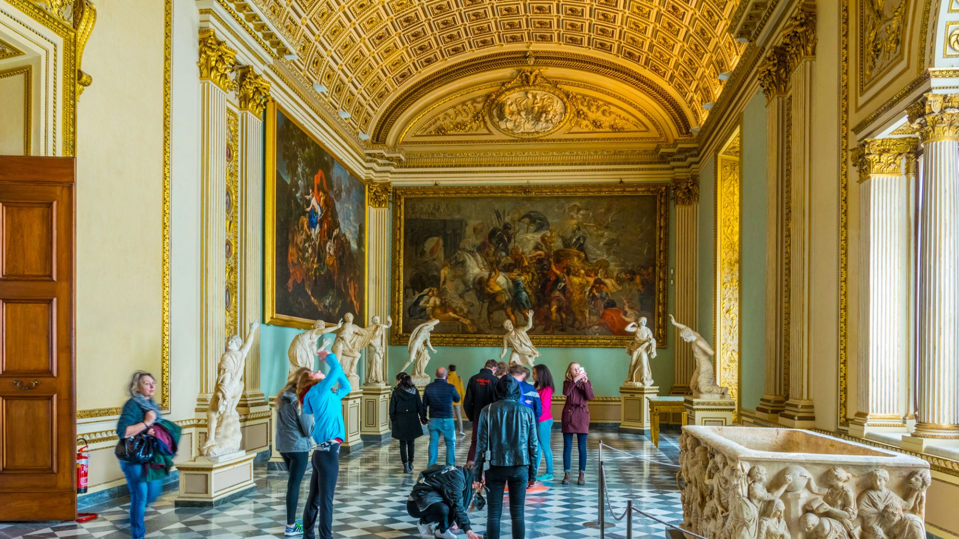 I visitatori esplorano una galleria museale ricca di statue di marmo, dipinti classici, dettagli dorati, alte colonne e un pavimento a motivi sotto un soffitto decorativo a volta.
