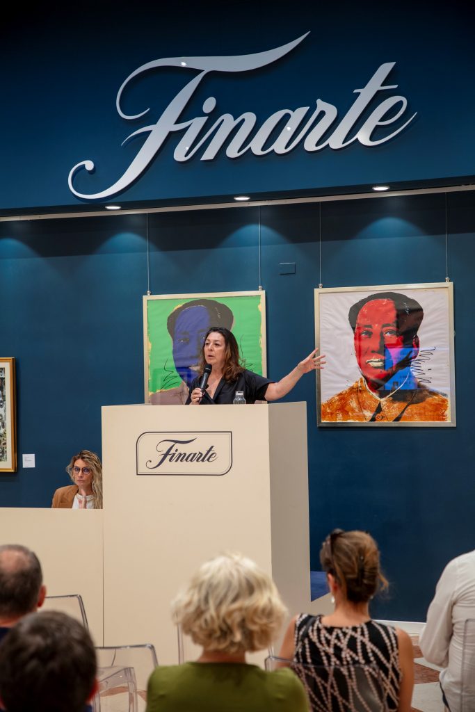 Una donna parla su un podio con la scritta "Finarte" durante un'asta, con quadri Bertinotti colorati di un uomo alle sue spalle e un'altra donna seduta vicino.