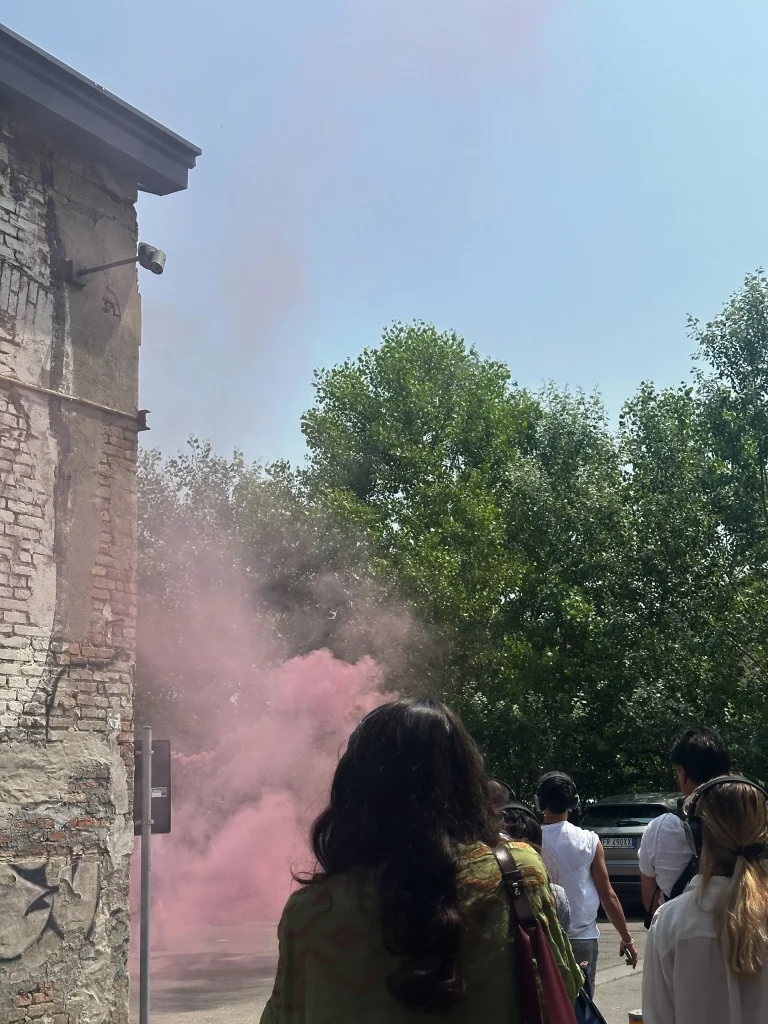 Un gruppo di persone cammina all'aperto vicino a un edificio di mattoni consumato dalle intemperie, con un fumo rosa che si diffonde nell'aria come in una scena di Santa di Maurizio Cattelan e alberi verdi visibili sullo sfondo in una giornata di sole.