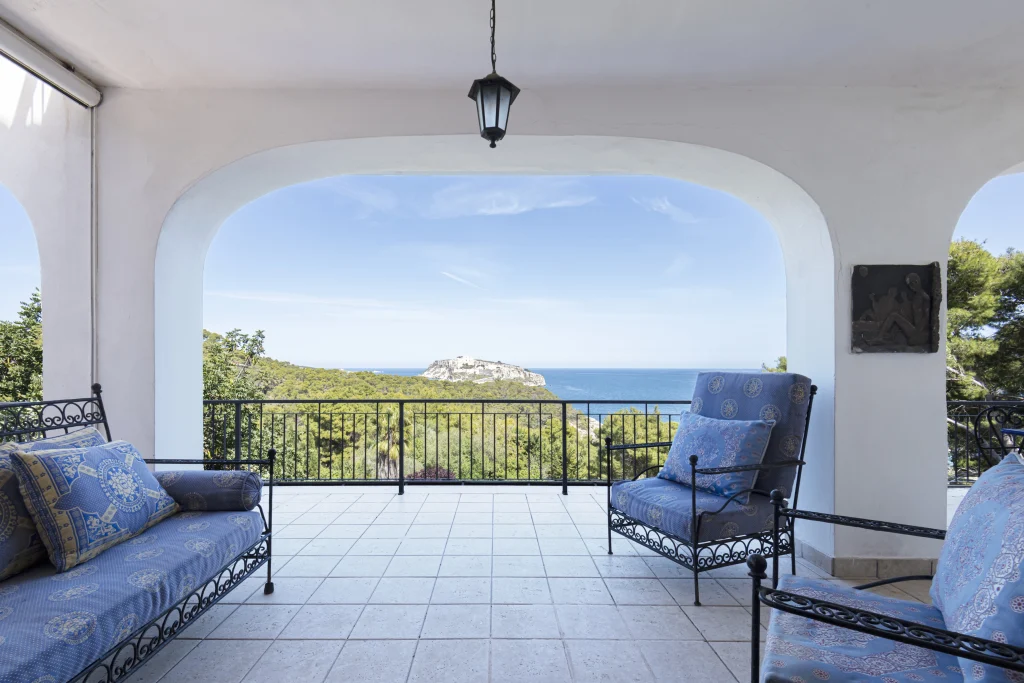 Un patio coperto con mobili in ferro battuto e cuscini blu si affaccia su una vegetazione lussureggiante e su una vista lontana del mare e di una piccola isola, che ricorda Lucio Dalla Tremiti, sotto un cielo limpido e azzurro.