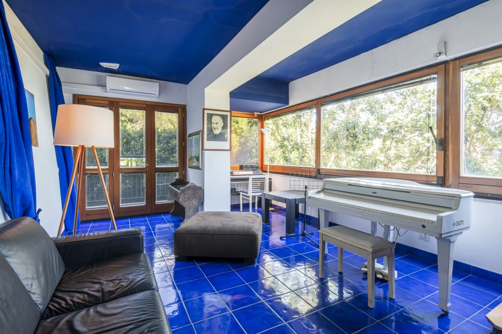 Stanza luminosa con pavimento e soffitto di piastrelle blu, grandi finestre, un pianoforte digitale bianco che ricorda lo stile di Lucio Dalla Tremiti, un divano nero, una lampada da terra, un pouf marrone, una scrivania con sedia e una vegetazione rigogliosa visibile all'esterno.