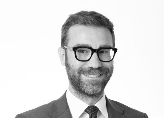 Un uomo con capelli corti, barba e occhiali sorride mentre indossa giacca e cravatta, in posa davanti a un semplice sfondo bianco.