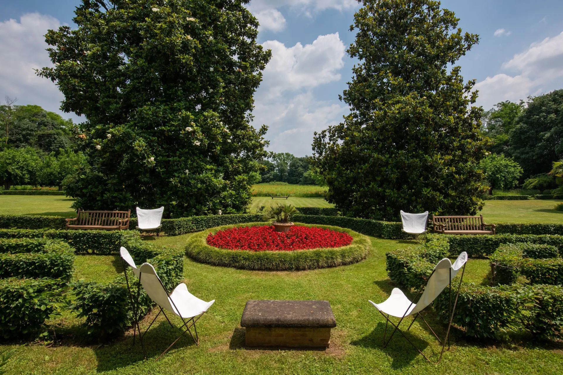 Un giardino formale che ricorda una villa di Lina Wertmüller, con due panchine, quattro sedie bianche a farfalla, un tavolo in pietra e un'aiuola circolare con fiori rossi tra siepi curate e alberi alti sotto un cielo parzialmente nuvoloso.