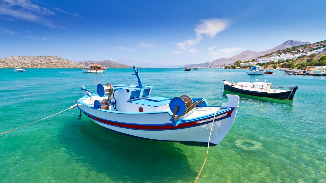 Una piccola barca da pesca bianca e blu galleggia sull'acqua limpida e turchese, legata con una corda: una scena idilliaca che suggerisce la vita tranquilla di una pensione estera. Un'altra barca e una costa collinare con edifici appaiono sullo sfondo di un cielo blu brillante.