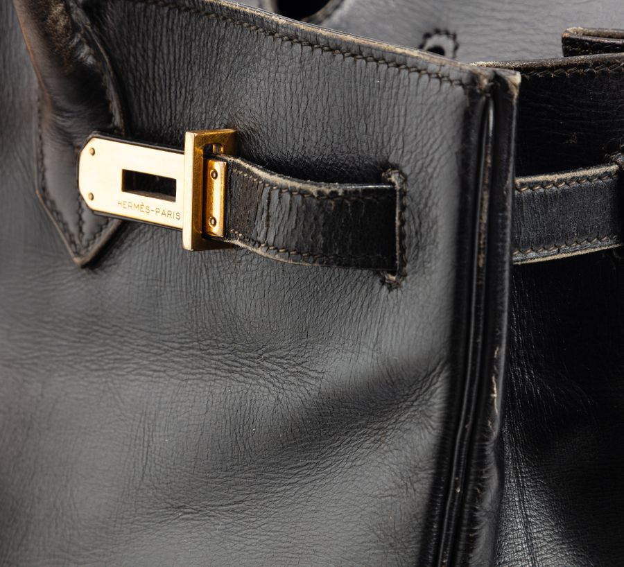 Primo piano di una borsa Jane Birkin in pelle nera con chiusura dorata con inciso "HERMÈS - PARIS". La texture della pelle e le cuciture dettagliate sono chiaramente visibili.