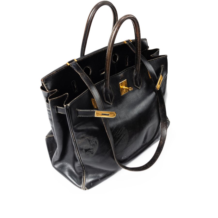 Una borsa Jane Birkin in pelle nera con finiture dorate, due manici e una forma strutturata è mostrata da una vista angolata dall'alto su uno sfondo bianco. La borsa appare leggermente usurata con pieghe e graffi visibili.