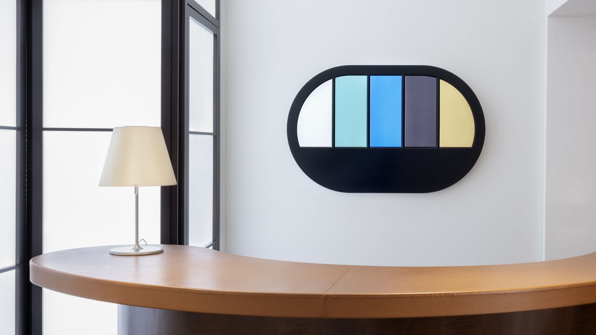 Un'area reception minimalista con una scrivania in legno curvato, una lampada da tavolo bianca e un'opera d'arte moderna a parete della collezione Julius Baer arte, caratterizzata da cinque pannelli verticali colorati incorniciati in una forma ovale nera.