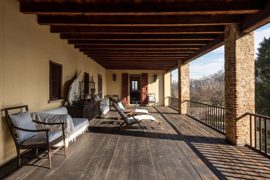 Una spaziosa veranda coperta in legno ispirata a una villa di Lina Wertmüller, con travi rustiche, colonne in pietra, sedie a sdraio e divani; la luce del sole proietta ombre attraverso la ringhiera, con vista sugli alberi sullo sfondo.