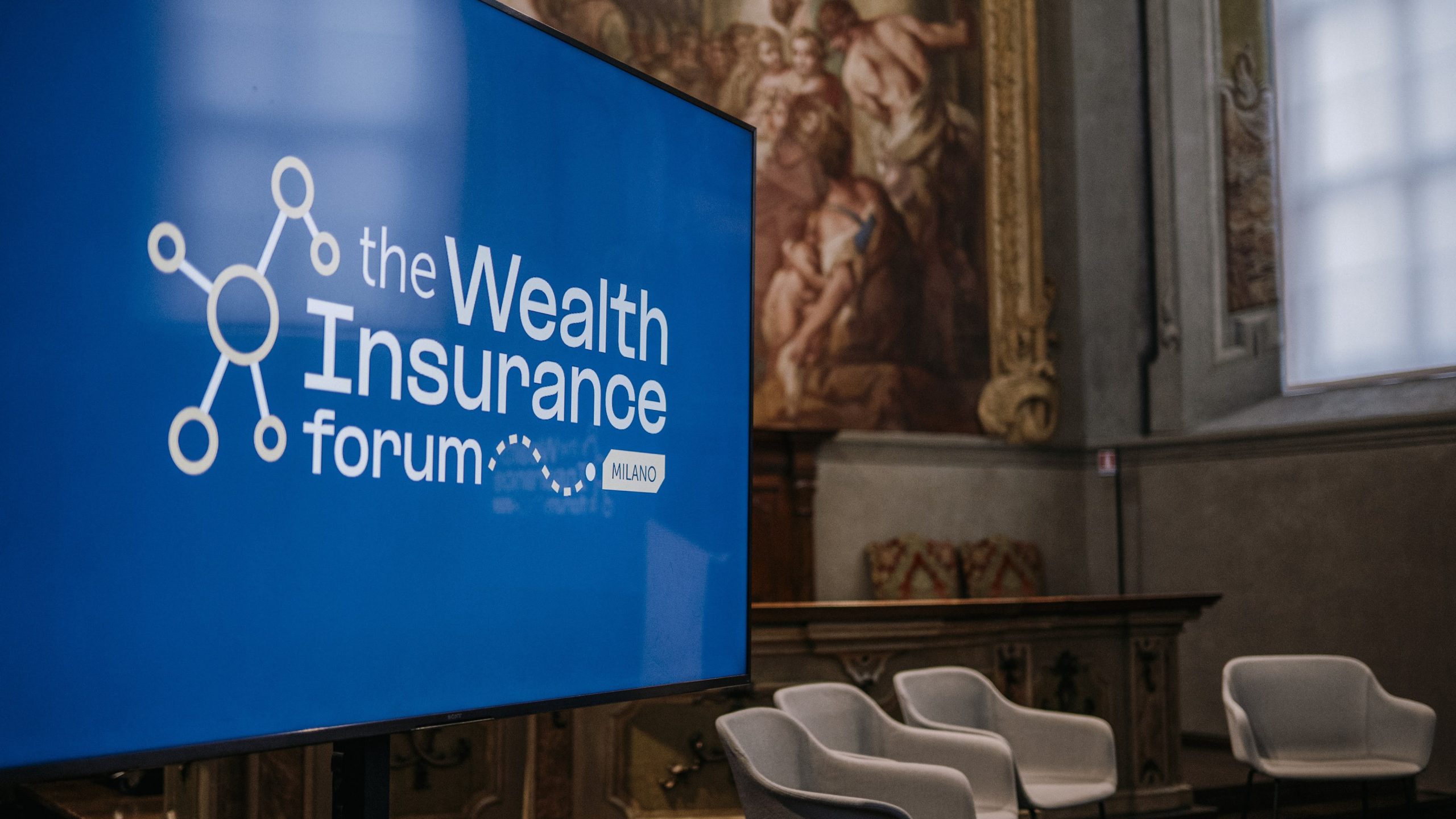 Uno schermo televisivo mostra "il Wealth Insurance forum MILANO" in una sala con pareti decorate e sedie bianche vuote disposte per una tavola rotonda. La luce naturale entra da una grande finestra.
