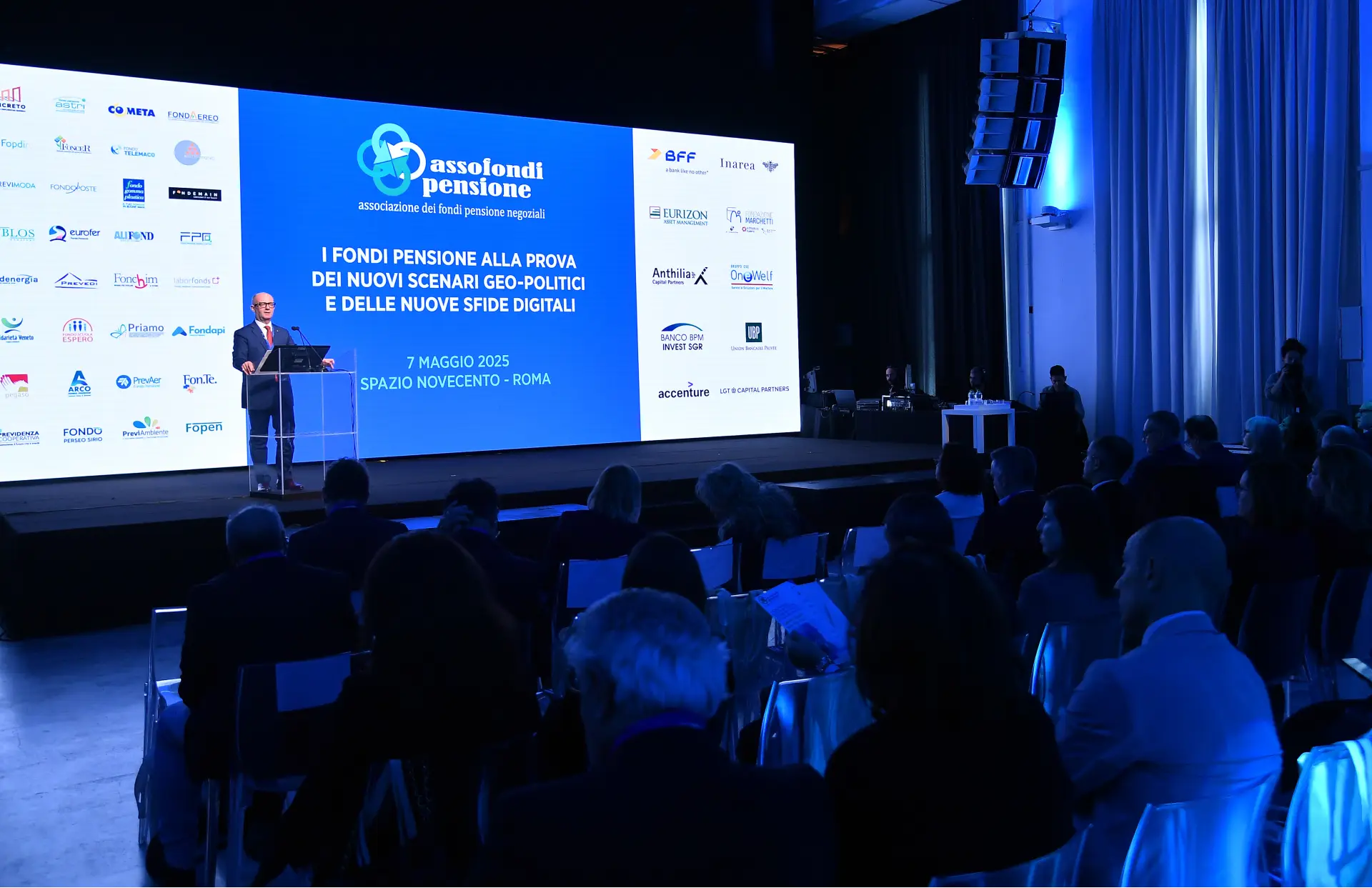 Un oratore è in piedi su un podio davanti a un grande schermo durante una conferenza e presenta a un pubblico seduto in una sala poco illuminata. Sullo schermo vengono visualizzate le informazioni sull'evento e i loghi degli sponsor.