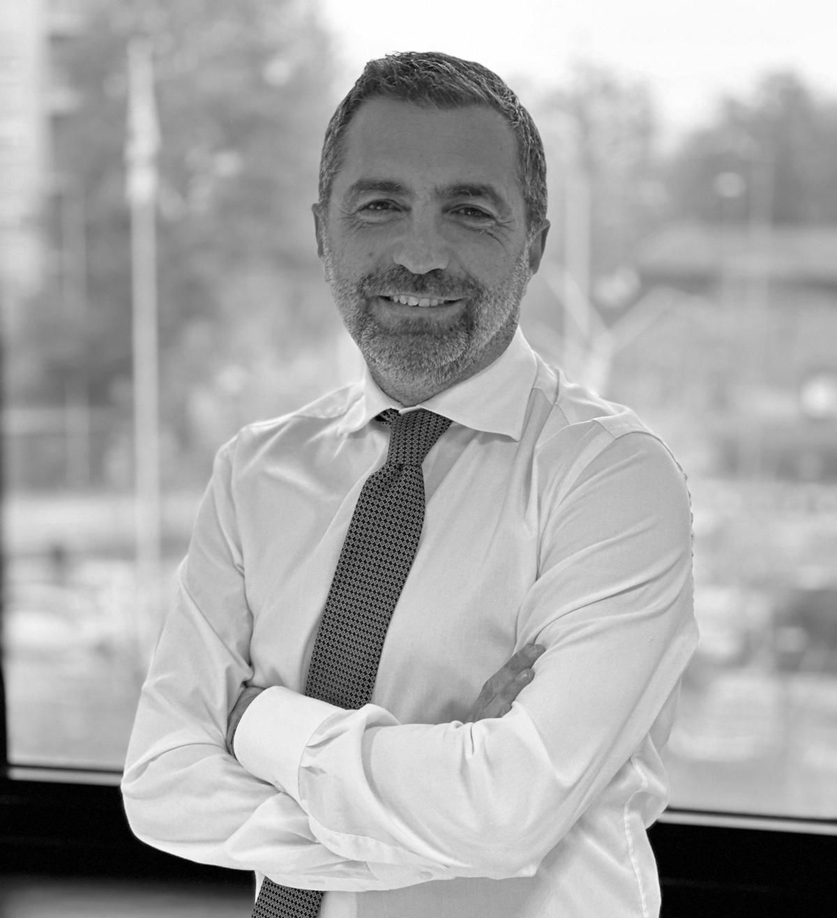Un uomo con capelli corti e barba, vestito con camicia e cravatta, sta in casa con le braccia incrociate e sorride all'obiettivo. Lo sfondo mostra una finestra con una scena all'aperto in soft focus, evocando un'atmosfera tranquilla da decio desio.