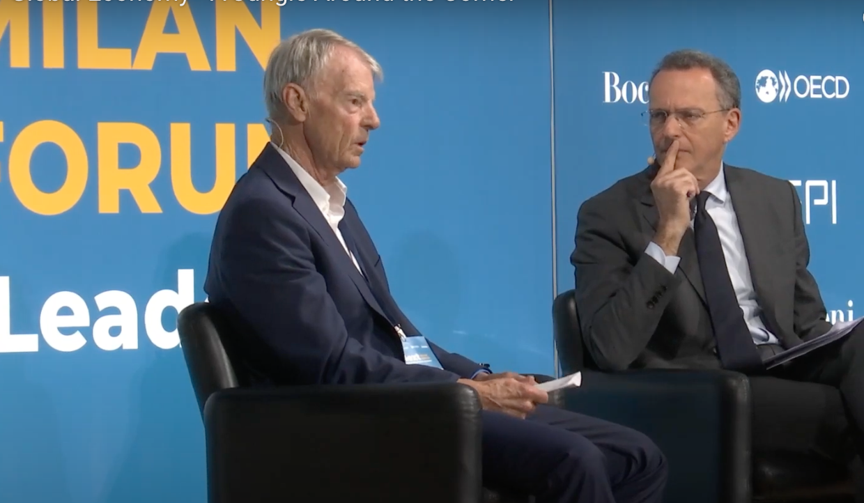 Due uomini in giacca e cravatta siedono su sedie nere sul palco e discutono. Sullo sfondo blu alle loro spalle compaiono testi gialli e bianchi, tra cui "FORUM DI MILANO" e loghi come "OCSE".