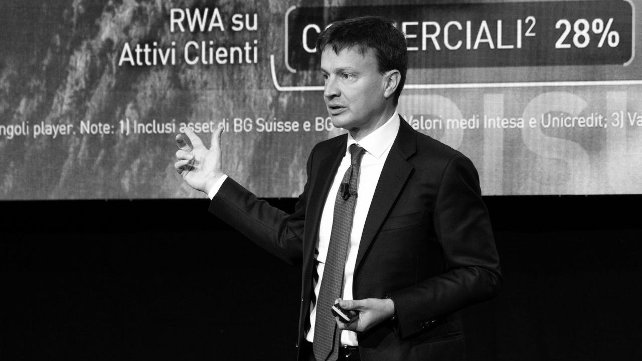 Un uomo in giacca e cravatta fa una presentazione sul palco, gesticolando con una mano. Alle sue spalle c'è un grande schermo con testi in italiano e termini finanziari. L'immagine è in bianco e nero.