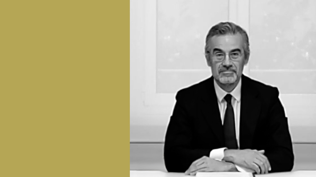 Un uomo in giacca e cravatta siede a una scrivania con le mani conserte, davanti a una finestra. La metà sinistra dell'immagine è riempita da un colore giallo senape pieno. La foto è in bianco e nero.