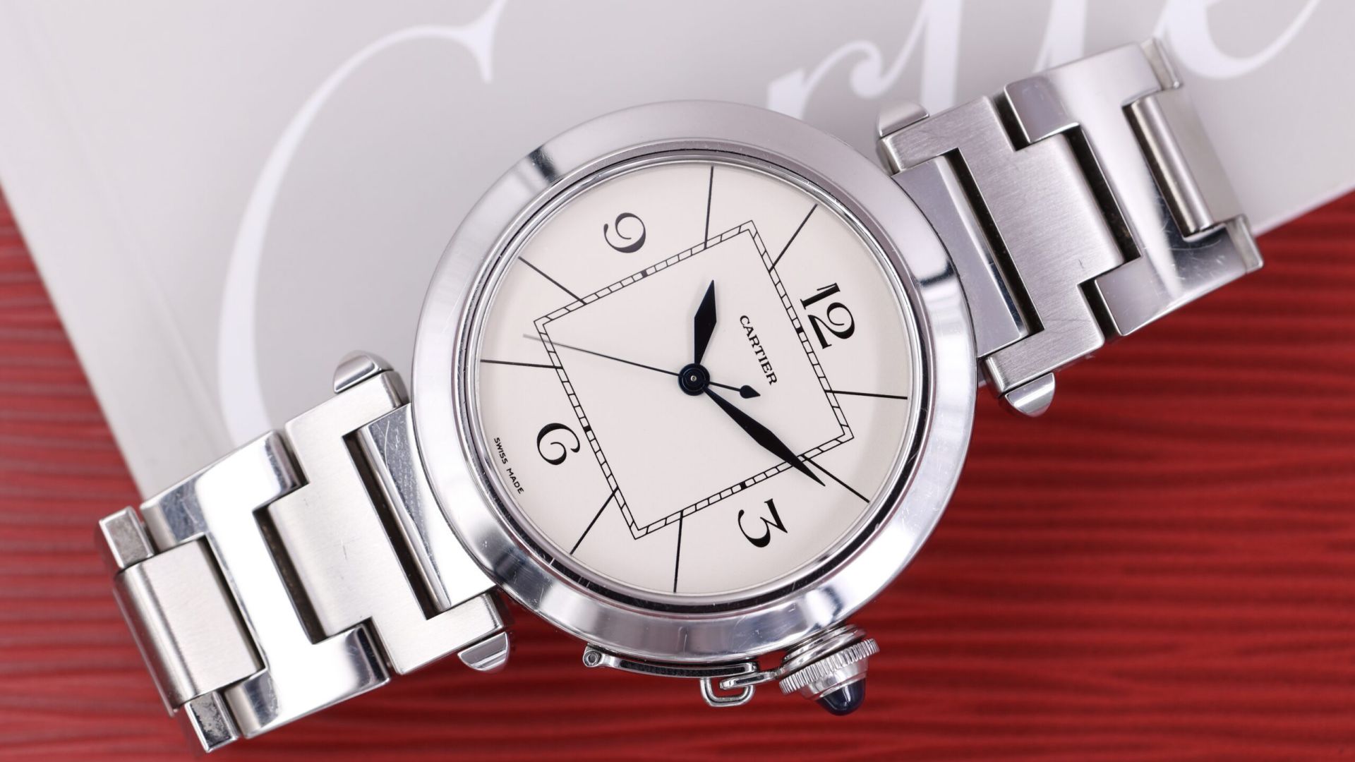 Un orologio da polso Cartier in argento con quadrante rotondo bianco, numeri neri, lancette blu e cinturino a maglie metalliche è esposto su una superficie rossa e bianca: una scelta elegante tra gli orologi di proprietà.