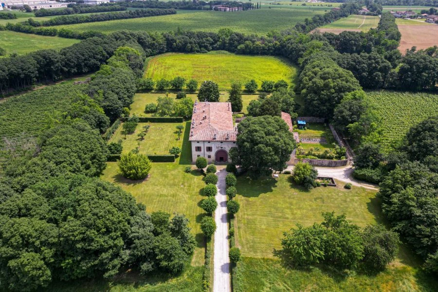 Veduta aerea di una grande villa di Lina Wertmüller con tetto in tegole rosse, circondata da giardini e alberi, immersa nel verde dei campi e dei terreni agricoli, con un lungo viale che conduce all'elegante ingresso.