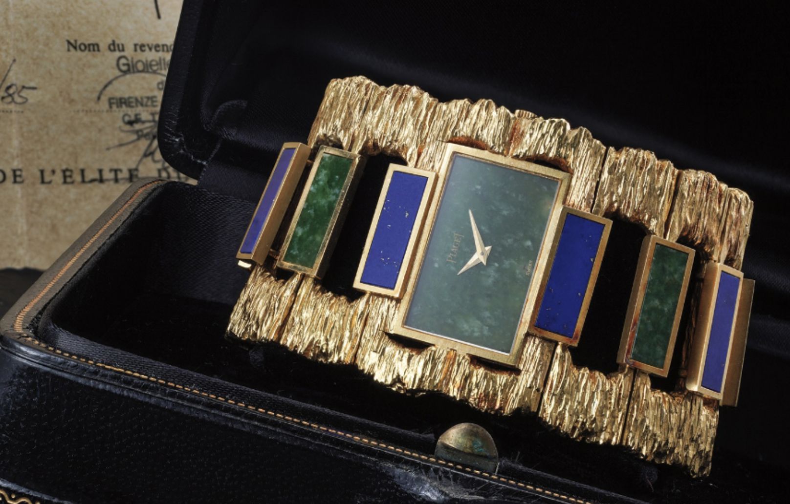 Lussuoso orologio da polso orologi gioiello in oro con cinturino testurizzato, con intarsi rettangolari di pietre verdi e blu, esposto in una cassa nera aperta con documenti sullo sfondo.