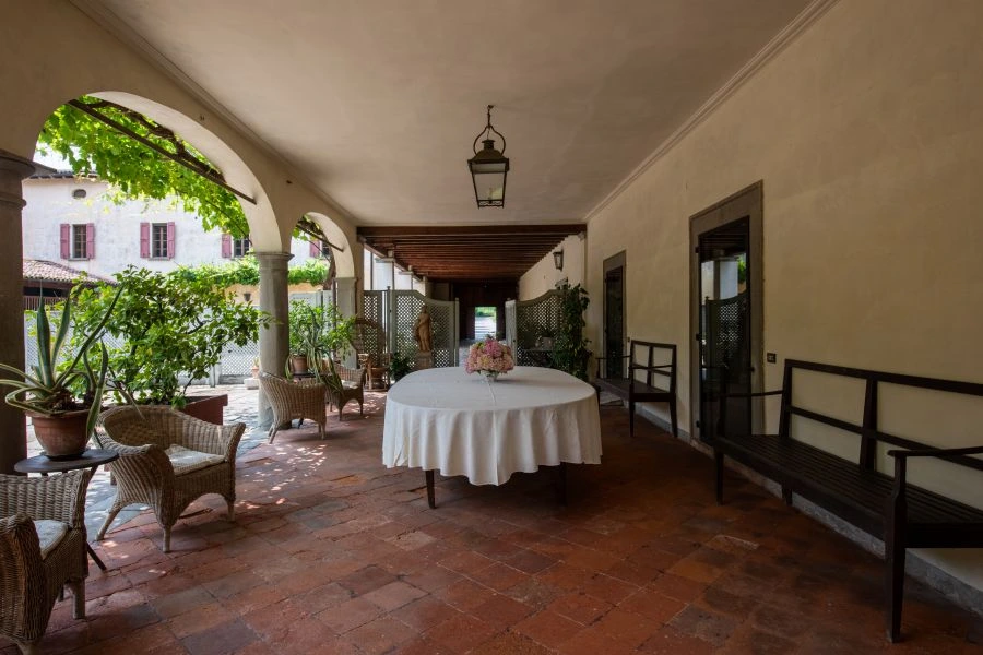Il patio coperto di una villa di Lina Wertmüller è caratterizzato da un tavolo rotondo drappeggiato di bianco, circondato da sedie di vimini, panche e piante in vaso. Le aperture ad arco lasciano intravedere la luce del sole e il verde all'esterno, creando un'atmosfera tranquilla e invitante.