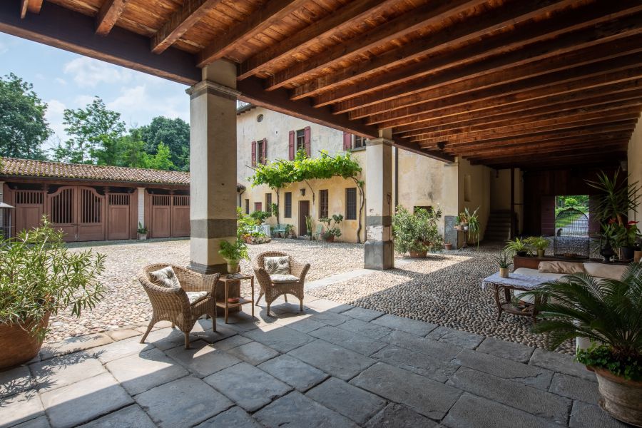 Un cortile rustico con pavimento in pietra, sedie in vimini, piante in vaso e un tetto con travi in legno rievoca il fascino di una villa di Lina Wertmüller. Sullo sfondo ci sono stalle, un albero e un edificio coperto di viti con finestre.