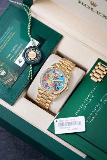 Un orologio Rolex Puzzle d'oro con un quadrante colorato è esposto in una scatola verde aperta insieme alla scheda di certificazione, alle etichette, al libretto di garanzia e a una maglia extra del bracciale d'oro.