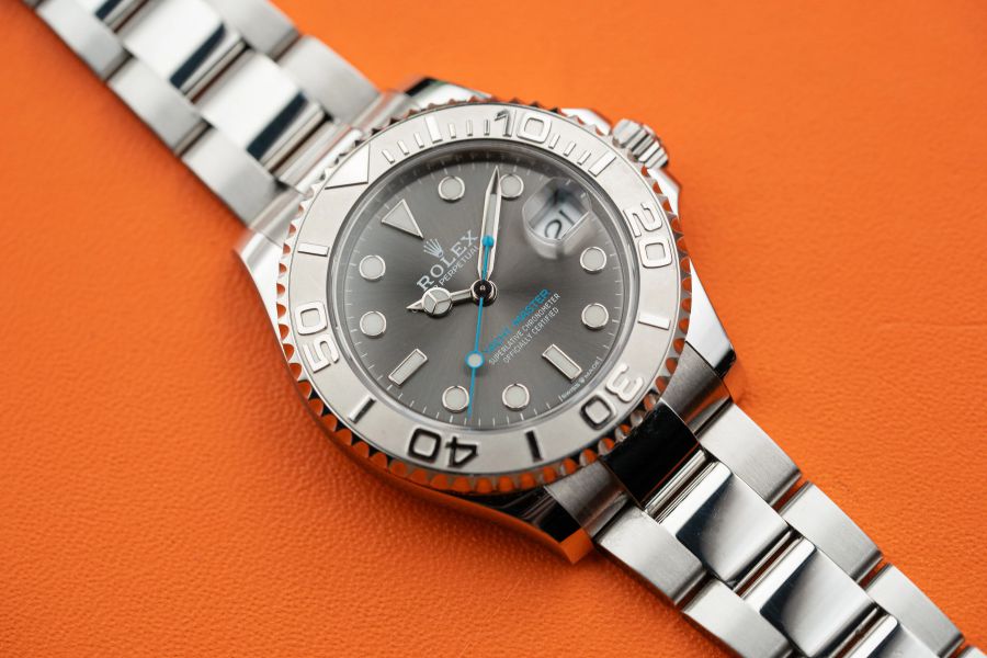 Un orologio Rolex Yacht-Master argentato con quadrante grigio, lunetta argentata e cinturino in metallo argentato è visualizzato su uno sfondo arancione: un accessorio orologiero ideale per un look estivo alla moda.