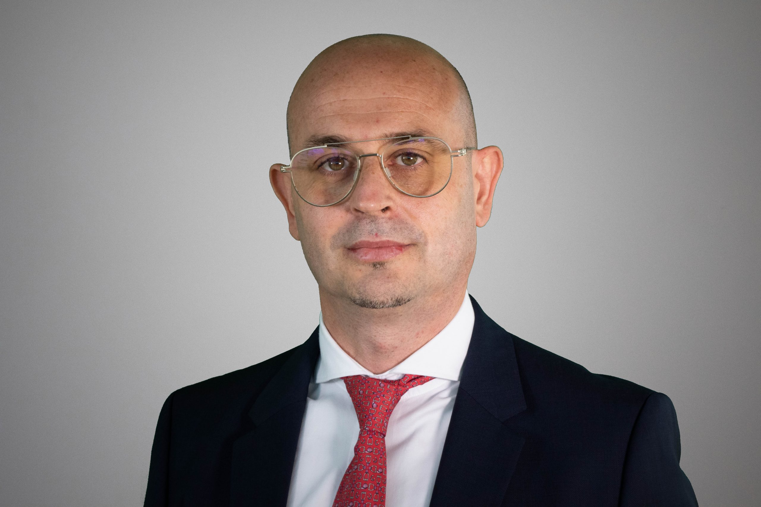 Un uomo calvo che indossa occhiali, abito scuro, camicia bianca e cravatta a motivi rossi si trova di fronte a uno sfondo grigio semplice e guarda direttamente la telecamera con un'espressione neutra.