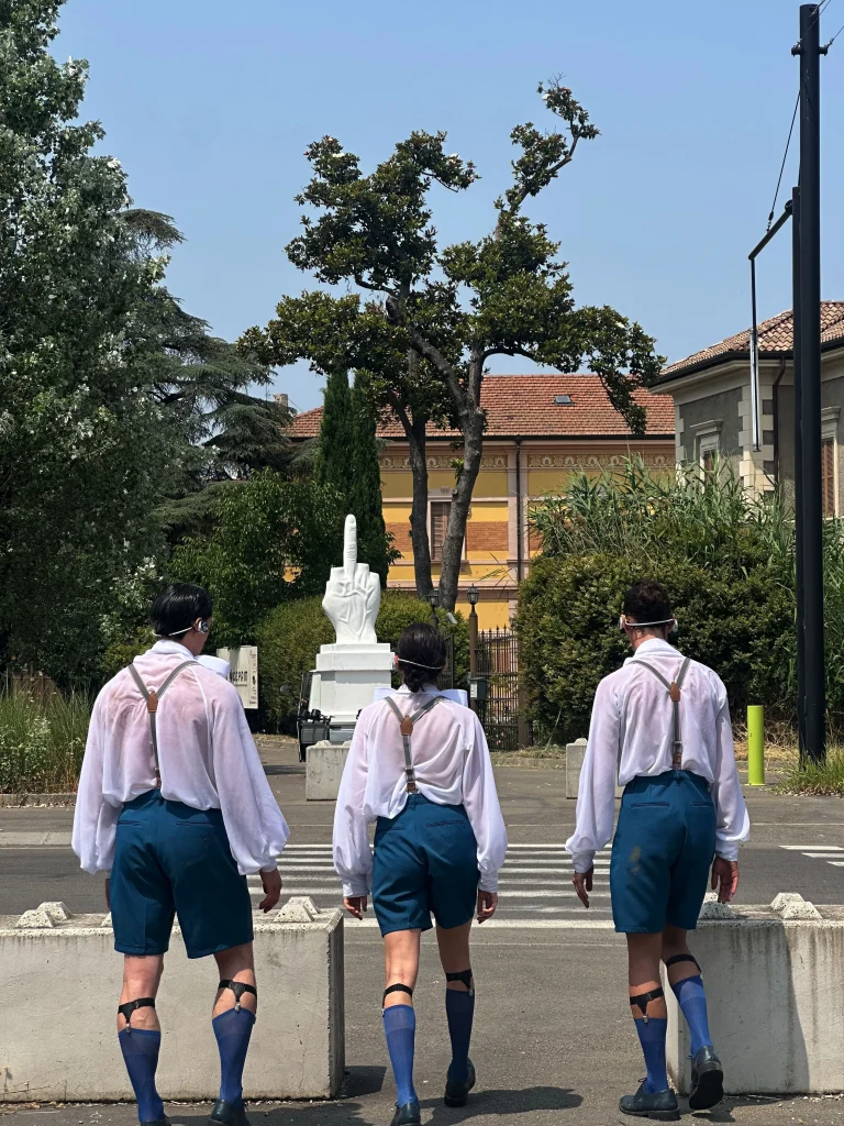 Tre persone in abiti coordinati camminano verso un passaggio pedonale, dove si trova il Babbo Natale di Maurizio Cattelan, una grande scultura bianca di una mano che mostra il dito medio. Alberi ed edifici incorniciano l'audace opera d'arte sullo sfondo.