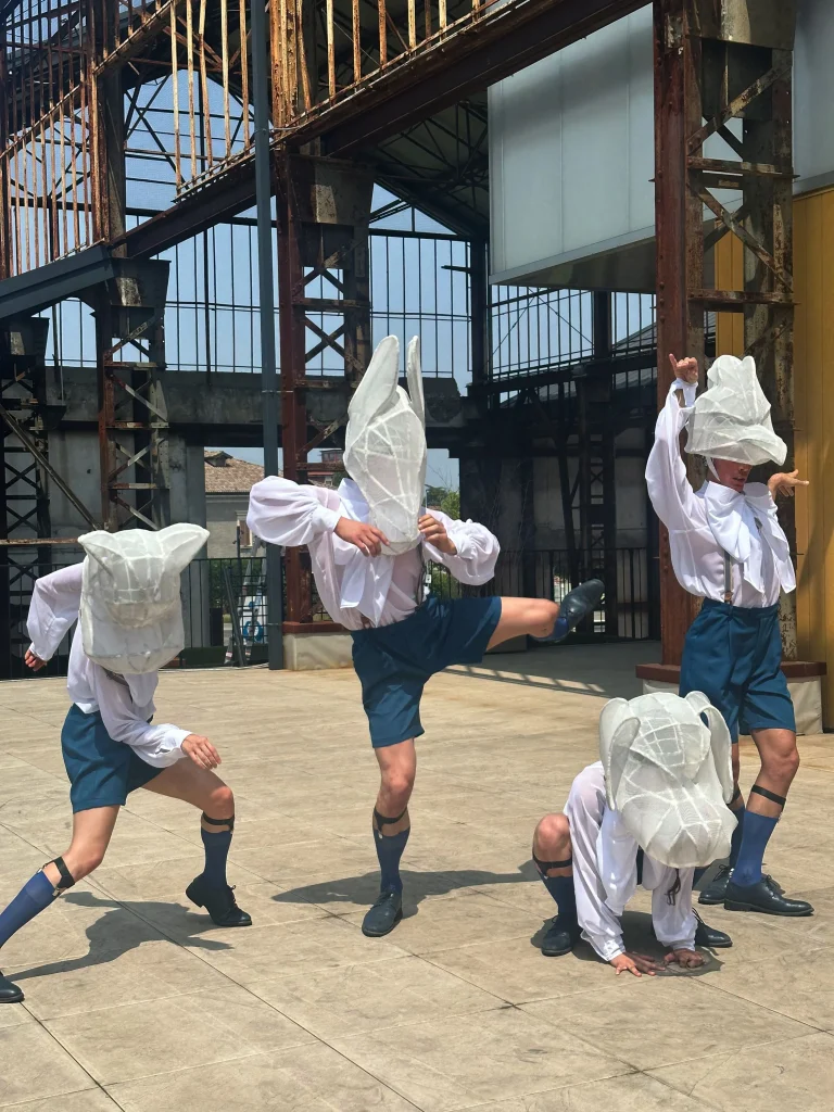 Quattro performer in pantaloncini blu, camicie bianche e calzettoni coordinati posano energicamente all'aperto. Ognuno di loro indossa un grande copricapo astratto bianco che ricorda teste di animali o insetti, in uno stile surreale che evoca Maurizio Cattelan Santa.
