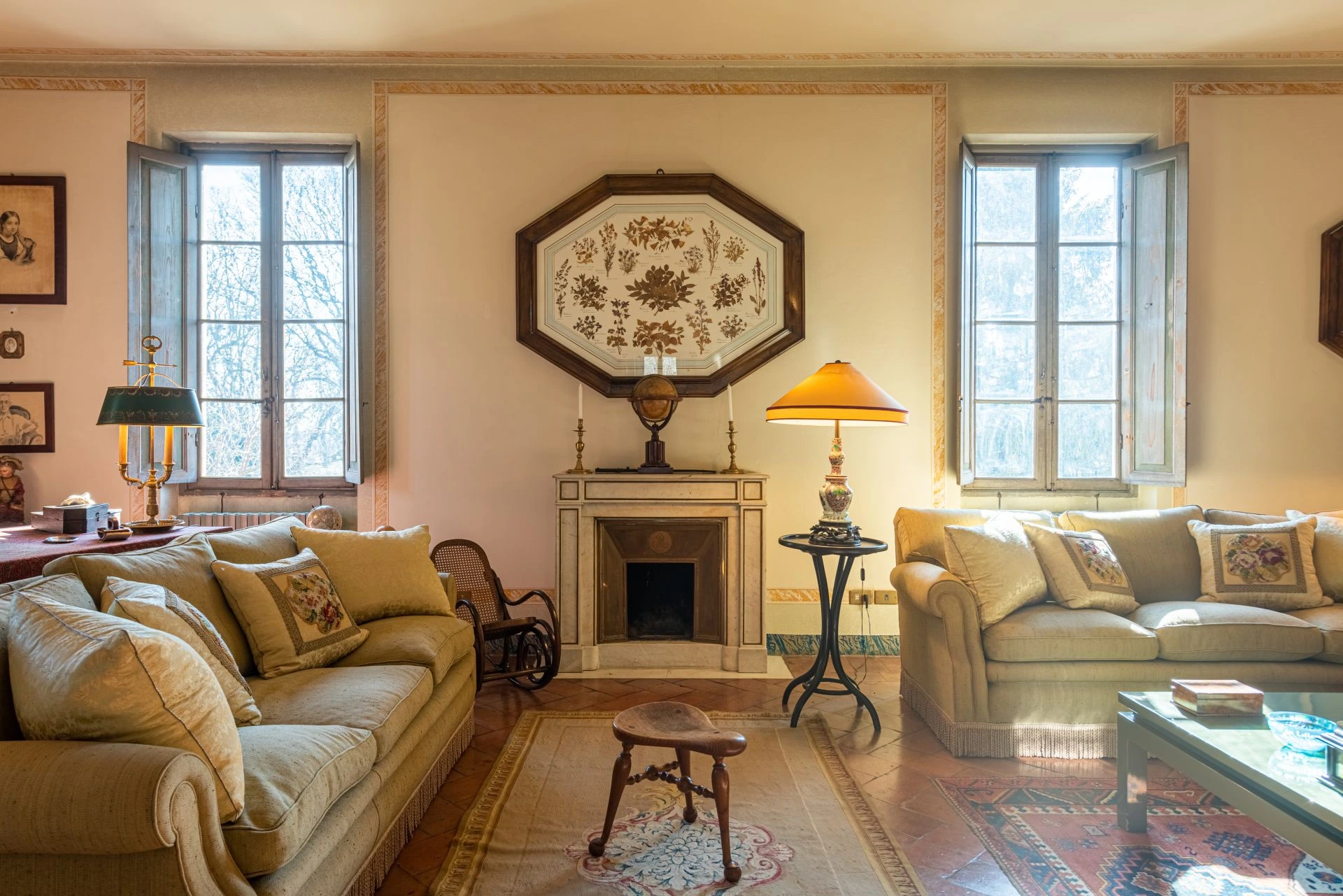 Un accogliente soggiorno che ricorda una villa di Lina Wertmüller, con due divani beige, un camino coronato da opere d'arte, grandi finestre che riempiono lo spazio di luce naturale e un arredamento che crea un'atmosfera calda e invitante.