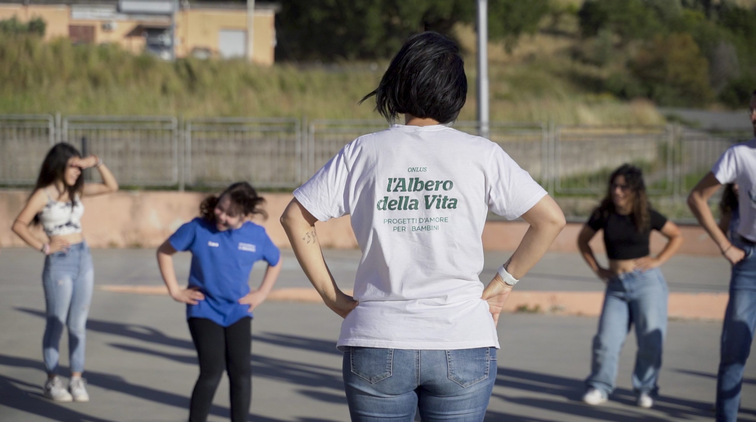 Un gruppo di persone si trova all'aperto con le mani sui fianchi. L'attenzione è rivolta a una donna con una maglietta bianca con la scritta "L'Albero della Vita" stampata sul retro, che guida gli altri in attività che promuovono la rinascita sociale e il legame con la comunità.