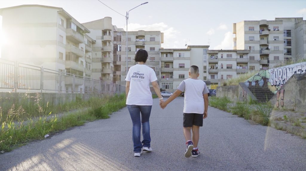 Una donna e un ragazzo che si tengono per mano camminano lungo una strada urbana deserta, fiancheggiata da condomini, inondata dalla luce del sole. Le loro camicie bianche spiccano, quella della donna è caratterizzata da una scritta sul retro, simbolo silenzioso di rinascita sociale.
