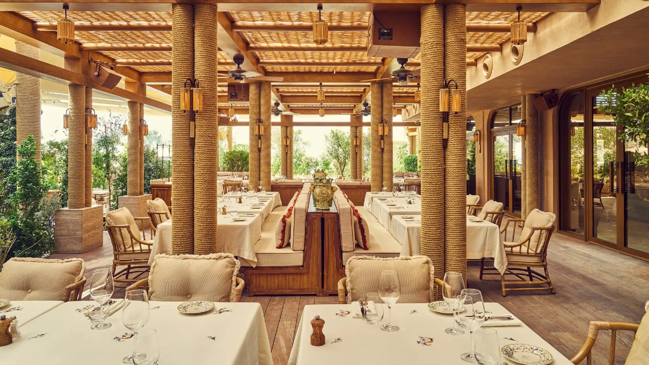 Elegante ristorante di Dubai con colonne in legno, sedie imbottite e tovaglie bianche. Le grandi finestre e il verde creano un'atmosfera luminosa e ariosa. L'illuminazione calda e l'arredamento naturale conferiscono un'atmosfera accogliente e invitante. I tavoli sono apparecchiati per gli ospiti.