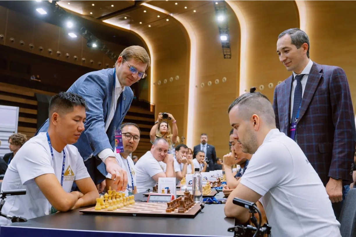 Un uomo in abito blu fa la mossa di apertura su una scacchiera tra due giocatori seduti in camicia bianca, osservati da diverse persone in una sala da torneo moderna e ben illuminata.